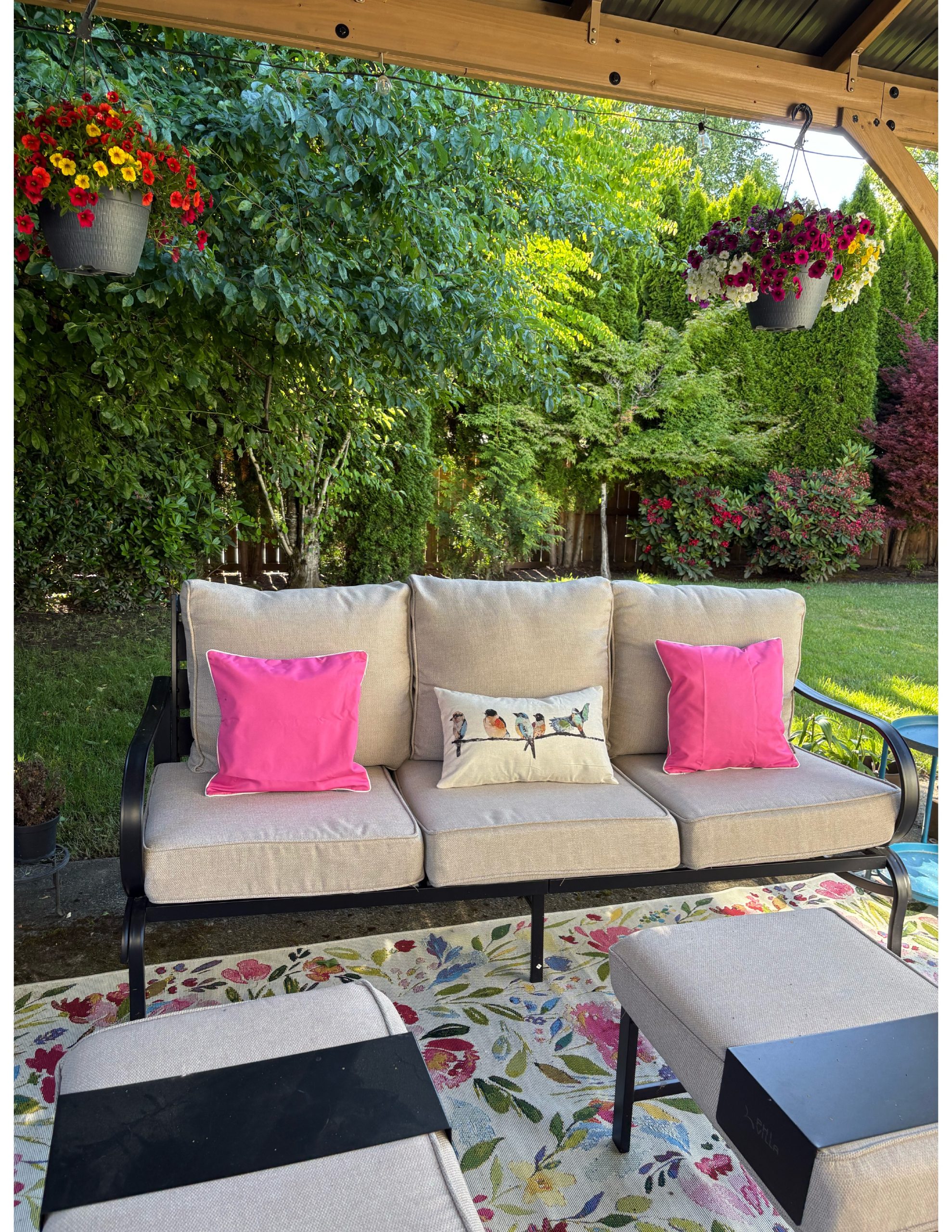 Backyard patio with cushioned outdoor seating, colorful accent pillows, a decorative bird pillow, hanging flower baskets, and mature landscaping.